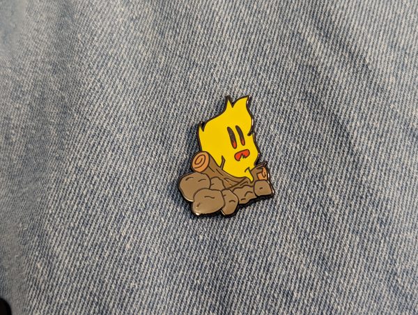 Campfire Hard enamel pin fitted to denim. Yellow fire, burning on wood logs and rocks.