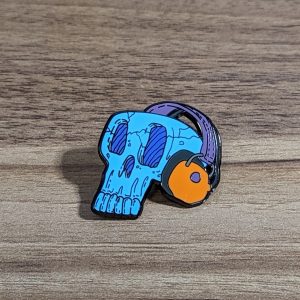 Blue Skull wearing headphones hard enamel pin. The headphones are orange with a purple strap.