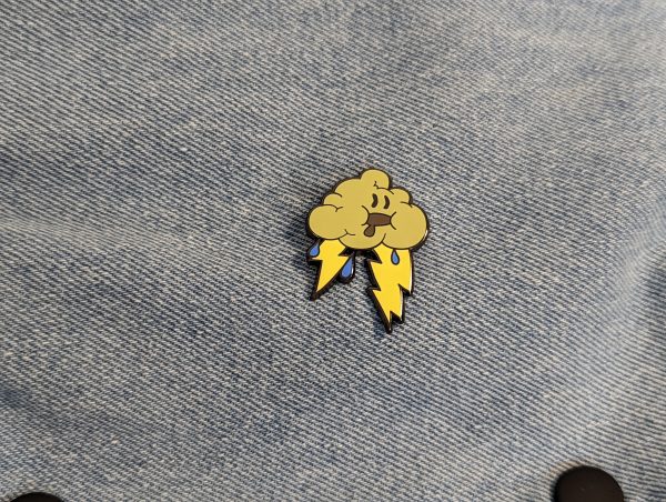 The storm cloud hard enamel pin is grey with yellow lightning bolts and blue rain drops. The pin has a drooling face!